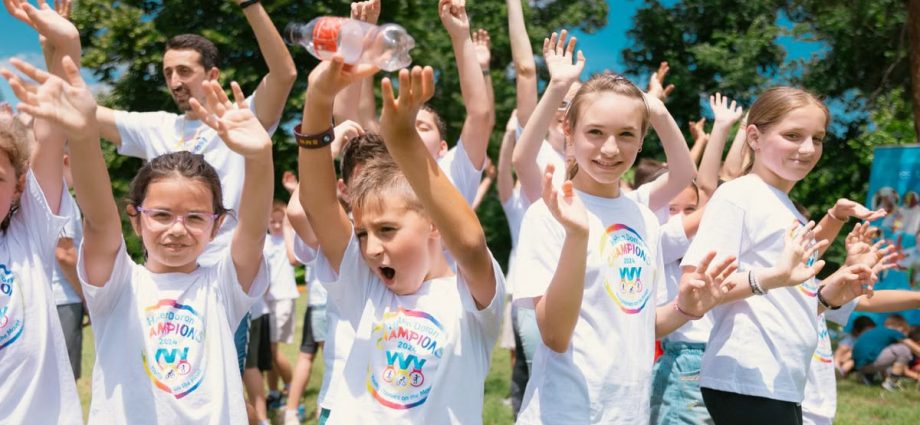 Group of cheerful children raising hands in a park during a sunny day, wearing t-shirts with event logos.